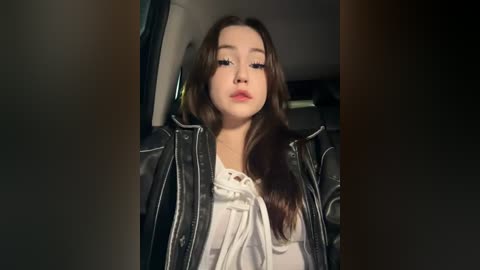 Media: Video of a young woman with long brown hair, fair skin, and a neutral expression, wearing a white blouse and black leather jacket, standing in a dimly lit room with blurred background.