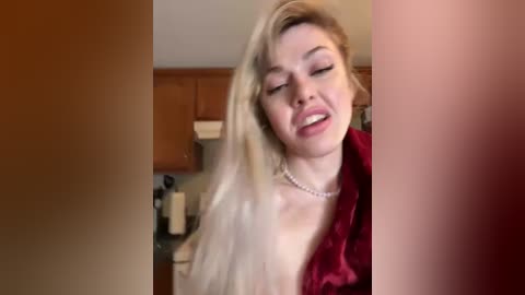 Media: A candid video shows a blonde woman with fair skin and a medium build, wearing a red robe, leaning against a kitchen counter, eyes closed, smiling. Wooden cabinets and kitchen items are visible in the background.