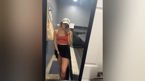 Media: Video of a woman in a gym changing room, wearing a red sports bra, black shorts, and a white hat, holding a phone, capturing her reflection in a mirror.