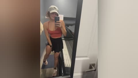 Media: Video of a woman in a gym, wearing a white bucket hat, red sports bra, black shorts, and sneakers, taking a selfie in a mirror.