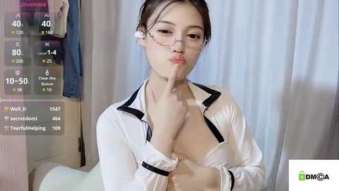 Media: Video of an Asian woman with glasses and a white blouse, making a kiss gesture. Background shows a room with clothes and a digital screen displaying statistics.