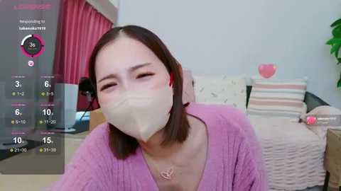 Media: Video of a young woman with straight, shoulder-length brown hair, wearing a pink sweater and a surgical mask, smiling. Background features a cozy bedroom with pink curtains and a bed with striped pillows.