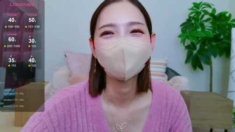 Media: Video of an East Asian woman with light skin and straight brown hair, wearing a pink sweater, beige mask, and gold earrings, sitting indoors, with a green plant in the background.