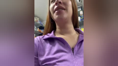 Media: Video of a woman with light skin and straight brown hair, smiling, wearing a purple zip-up hoodie, standing in a grocery store with shelves in the background.