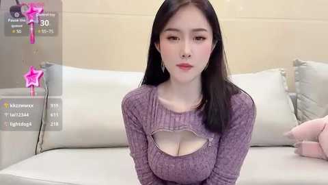 Media: Video of an East Asian woman with long black hair, fair skin, and a slender physique, wearing a purple ribbed sweater with a cut-out design revealing her ample cleavage. She sits on a white sofa in a minimalist room.