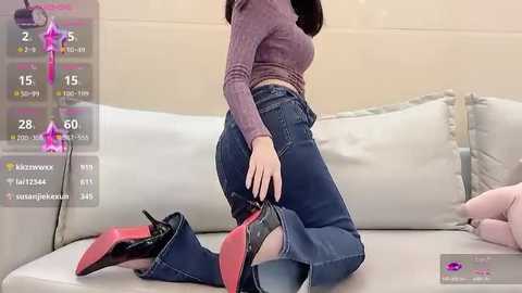 Media: A video of an Asian woman in a snug, long-sleeve purple top, high-waisted jeans, and black stiletto heels, kneeling on a beige sofa, adjusting her jeans.