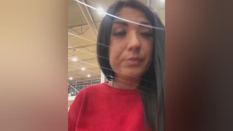 Media: Video of a young woman with long, dark hair and light skin, wearing a red sweater, standing in an indoor setting with a high ceiling and metal beams.