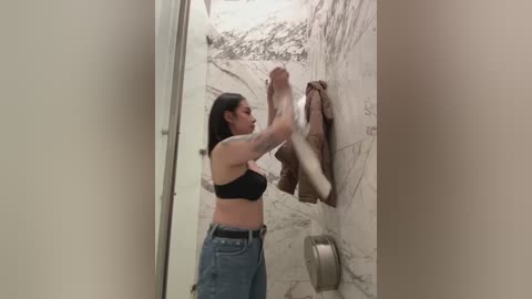 Media: A video of a woman with medium skin tone and dark hair, wearing a black sports bra and blue jeans, stretching her legs against a marble-tiled wall in a modern bathroom.