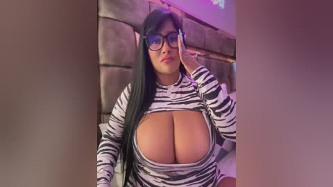 Media: Video of a voluptuous woman with long black hair, wearing a tight, zebra-print top that reveals her large breasts, standing indoors next to a brick wall.