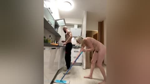 Media: Video of a nude, blonde woman cleaning a modern kitchen with a mop, while a shirtless, bald man in black pants observes.