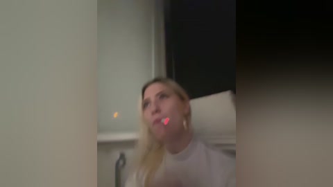 Media: Video of a blonde woman with a white shirt, blurred, mouth slightly open, in a dimly lit room with a black curtain and a window.