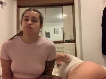 Media: Video of a young, light-skinned woman with a pouty expression, wearing a light pink top, sitting on a bed. A tattooed arm is visible. Background shows a white wardrobe with framed pictures and a mirror.