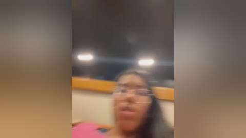 Media: Blurry video of a young woman with medium skin tone, possibly of African descent, wearing glasses and a pink top, sitting indoors with a blurred background of a car and dim lighting.