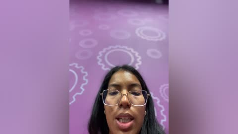 Media: Video of a woman with long black hair, wearing glasses, and a light purple top. Background features a purple wall with white circular patterns. She has a slightly open mouth, appearing to sing.