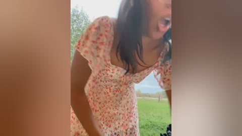 Media: A video of a woman in a floral dress bending over, with blurred background of trees and grass, capturing a candid moment outdoors.