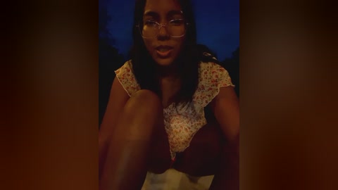 Media: A video of a dark-skinned woman with medium-length curly hair, wearing glasses, and a floral-patterned top, sitting on a bed with dim lighting.