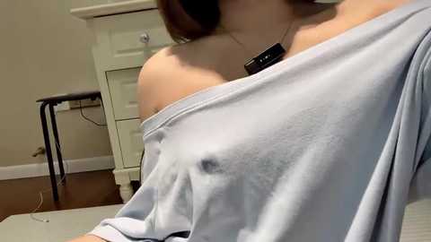 Media: Video of a light-skinned woman with auburn hair, wearing a loose, off-shoulder light blue sweater that exposes her left breast. Background includes a white dresser, black stool, and wooden floor.