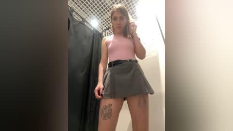 Media: Video of a young Caucasian woman with light skin and shoulder-length blonde hair, wearing a pink tank top and a gray pleated skirt. She poses in a bathroom with a black shower curtain and a grid-patterned ceiling.