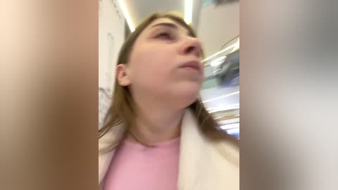 Media: A blurry video of a young woman with fair skin and long brown hair, wearing a pink shirt and white jacket, gazing upward in a subway car with fluorescent lighting and blurry passengers.