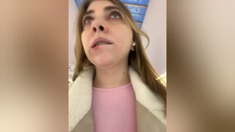 Media: Video of a Caucasian woman with long brown hair, wearing a pink shirt and beige jacket, looking upwards with a neutral expression.