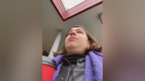 Media: Video of a woman with shoulder-length brown hair, wearing a purple jacket over a black zip-up hoodie, looking upward from a low angle, in a room with a red-framed skylight.