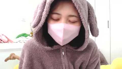 Media: A video of an Asian woman with short black hair wearing a pink face mask and a cozy, hooded brown bunny pajama set. The background is a white-walled bedroom with scattered clothes and a green towel.