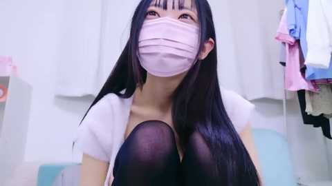 Media: Video of an Asian woman with long black hair, wearing a pink mask, white shirt, and black tights, sitting in a bright, minimalistic room with white walls and hanging clothes.