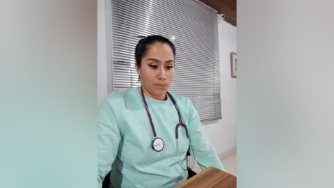 Media: Video of a Latina nurse in mint green scrubs, wearing a stethoscope, sitting at a wooden desk in a hospital room with white walls and horizontal blinds.