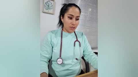 Media: Video of a Latina woman in mint-green scrubs, with a stethoscope around her neck, sitting at a desk in a medical office. Background includes a framed leaf design, a window with closed blinds, and a white wall.