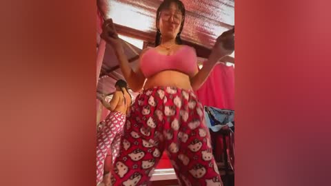 Media: Video of a young Asian woman in a pink sports bra and red, white, and pink Hello Kitty pajama pants, lifting weights in a rustic, red-lit room with wooden ceiling beams.