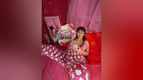 Media: Video of an Asian woman with glasses and black hair in a Hello Kitty pajama set, sleeping under a Hello Kitty duvet, surrounded by plush toys and red curtains in a cozy, pink-themed bedroom.