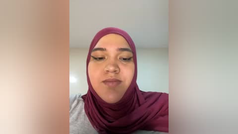 Media: Video of a light-skinned woman with full eyebrows, wearing a maroon hijab and a gray shirt, standing against a plain white wall.