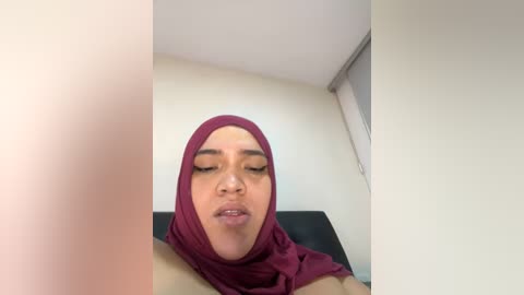 Media: A video of a young woman with light brown skin, wearing a maroon hijab and closed eyes, lying on a black couch in a plain room with white walls and a window.