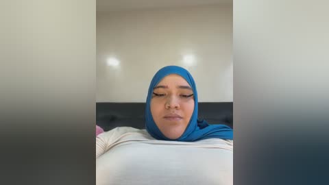 Media: A video of a woman with light brown skin, wearing a blue hijab and a white long-sleeved top, sitting on a black couch with a neutral background.