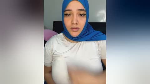 Media: Video of a young woman with medium skin tone, wearing a blue hijab and white t-shirt, sitting indoors. Background is blurred with a gradient of blue and purple.