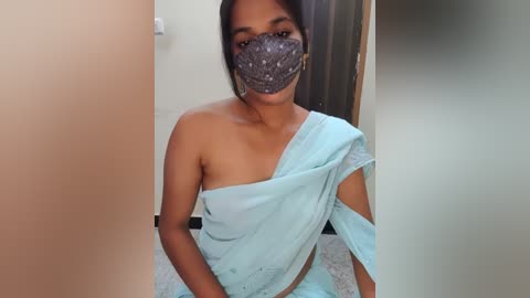 Media: Video of a South Asian woman with medium-brown skin and black hair, wearing a light blue saree and a gray mask, sitting on a beige floor, in a dimly lit room.