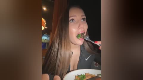 Media: Video of a young Asian woman with long, straight, dark brown hair, wearing a black Nike top, eating salad with a fork at a dimly lit restaurant.