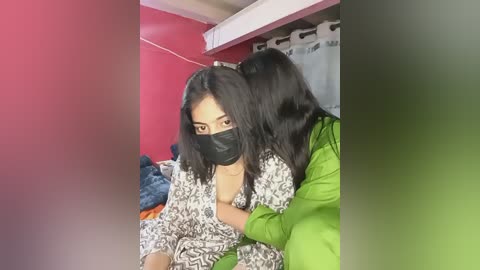 Media: Video of two women in a small, dimly lit room. One wears a black mask, a white and black patterned dress, and sits on a bed. The other, in a green jacket, embraces her. Background includes red walls, a blue blanket, and white curtains.