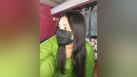 Media: A video of a woman with long black hair and a green jacket, wearing a black face mask, seated indoors with red and white walls, and a white ceiling fan visible.