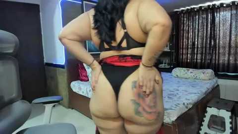 Media: Video of a curvy, dark-skinned woman with long black hair, wearing a black bra and thong, showing off her ample buttocks and a colorful tattoo on her right thigh. The room features a bed, curtains, and a chair.