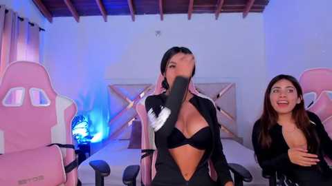 Media: Video of two young women in a gaming room with pink chairs and a blue light. One wears a black bra, the other a black top, both smiling.