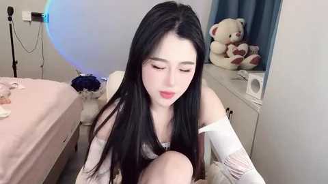 Media: Video of a pale, long-haired Asian woman with black hair, wearing a white off-shoulder top, sitting on a bed in a dimly lit room with blue curtains and a teddy bear.