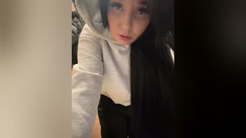 Media: A selfie of a young woman with long black hair, wearing a white hoodie and black skirt, taken in a dimly lit room.