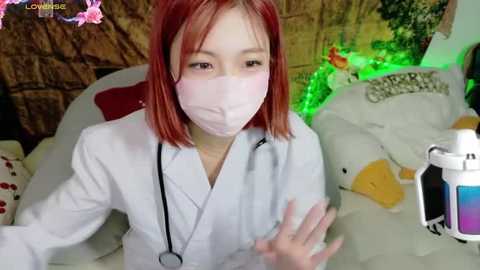 Media: Video of an Asian woman with shoulder-length red hair, wearing a white lab coat and face mask, holding a small device, against a background of a green light, plush toys, and a wooden wall.