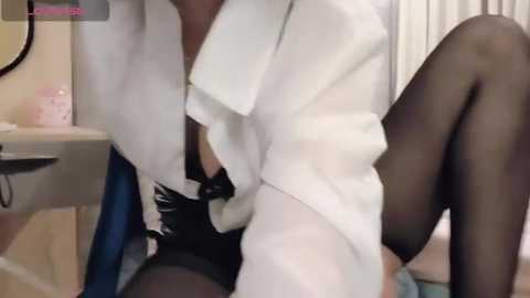 Media: Video of a person in a white lab coat with black gloves, sitting on a chair with black tights. Background includes a bathroom sink with toiletries and a window.