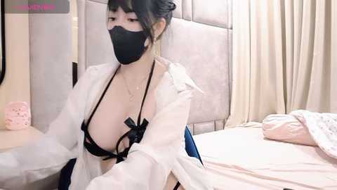 Media: Video of an East Asian woman with fair skin, wearing a black mask, revealing a black bra with a bow accentuating her large breasts. She has dark hair in a bun, white jacket, and is in a modern bedroom.