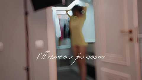 Media: A video of a woman in a tight, short yellow dress, posing in a mirror, with blurred background showing a closet. Text overlay reads, \"I'll start in a few minutes.\