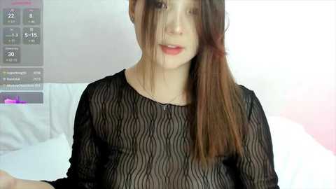 Media: A video of a young Asian woman with long brown hair, wearing a sheer black long-sleeve top, lying on a white bed. The background includes a digital screen displaying weather information.