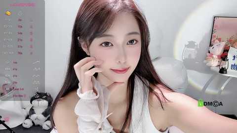 Media: Video of a young East Asian woman with long, straight black hair, fair skin, and light makeup, smiling softly. She's wearing a white sleeveless top. Background includes a calendar, desk, and a computer monitor.