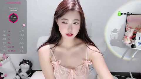 Media: Video of a young East Asian woman with long, straight dark hair and fair skin, wearing a pink lace bralette, sitting in a dimly lit room with a computer screen and stuffed animals.